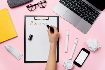Female hand writes resume with lipstick on white sheet of paper. Pink office desk with laptop,...
