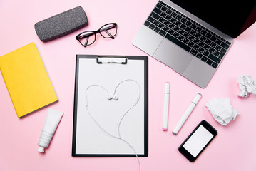 Woman's pink office desk with headphones laid out like a heart. Woman's workplace with laptop, smart phone with blank white screen, a cream, lipstick, eyeglasses and supplies.