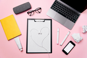 Woman's pink office desk with headphones laid out like a heart. Woman's workplace with laptop, smart phone with blank white screen, a cream, lipstick, eyeglasses and supplies.