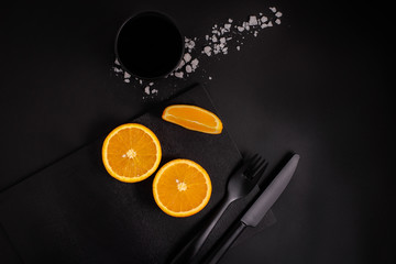 orange slice on black board with black glass and black cutlery on black background, top view, close up, copy space