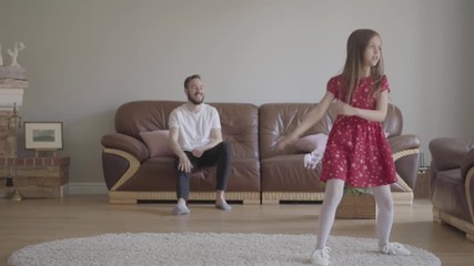 The little funny girl in red dress dancing in the foreground on the fluffy carpet. Bearded father sitting on the background on the leather sofa, clapping his hands and showing big finger