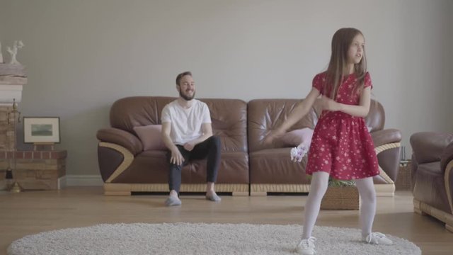 The little funny girl in red dress dancing in the foreground on the fluffy carpet. Bearded father sitting on the background on the leather sofa, clapping his hands and showing big finger