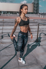 Girl athlete in tattoos, in summer in city, trains fitness, gymnastics workout. Sportswear leggings top. In the morning before the run. Background steps. It warms the muscles before exercise.