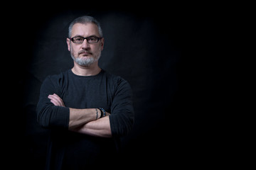 A man with a beard in a dark longsleeve with arms crossed strictly looking at the camera against a black wall