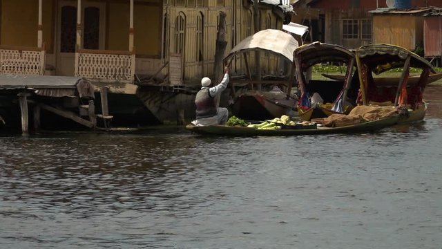 Wide shot of a man rowing a canoe full of vegetables in front of a houseboat and several docked shikara boats