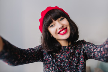 Charming brunette girl with bright make-up widely smiling while making selfie in her room. Close-up portrait of adorable young dark-haired woman wearing cute vintage outfit and red french beret