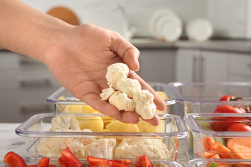 Woman putting cut cauliflower into box and containers with raw vegetables in kitchen, closeup
