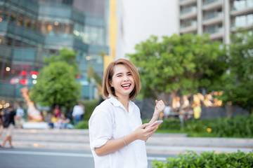 Young beautiful Asian lady celebrate with smart phone in the street in a sunny summer day, success happy pose, internet technology.