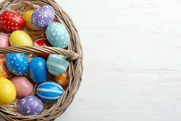 Wicker basket with painted Easter eggs on wooden table, top view. Space for text