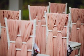 Beautiful setting for outdoors wedding ceremony. Chairs for guests of the bride and groom are decorated in the same style, decorated with fashionable fabric.  chairs are placed convolution with  rose 