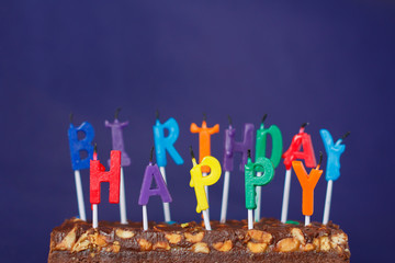 Happy Birthday Brownie Cake with Peanuts, Salted Caramel and Colorful Unlighted Candles on the Violet Background. Copy Space for Text.