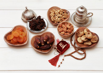 Ramadan iftar party concept. Islamic holy book Quran and rosary beads with delicious dates, dried figs, dried apricots, walnuts, almonds, raisins on bamboo plates on a white wooden table background.