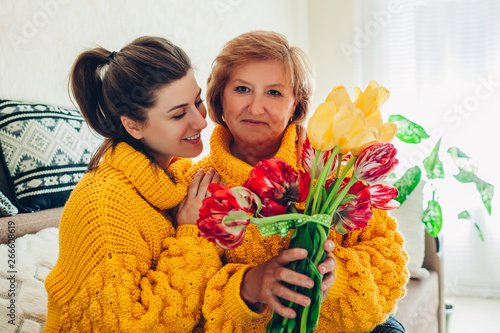 Daughter giving her mother bouquet of flowers at home as Mother's day present wearing similar sweaters