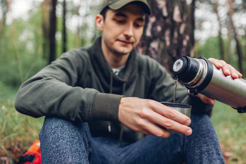 Man tourist pours hot tea out of thermos in spring forest. Camping, traveling and sport concept