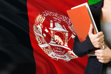 Learning Afghan language concept. Young woman standing with the Afghanistan flag in the background. Teacher holding books, orange blank book cover.