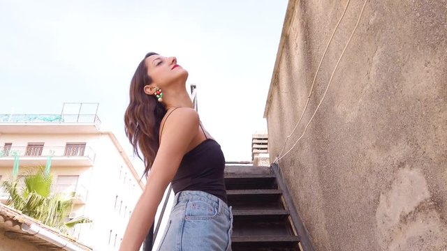 Giovane bella ragazza con capelli lunghi, jeans blu e vestito nero sta posando per foto di moda sulla scala all'esterno di casa vicino al terrazzo.