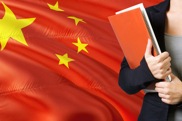 Learning Chinese language concept. Young woman standing with the China flag in the background. Teacher holding books, orange blank book cover.