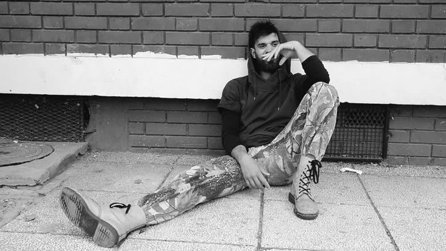Homeless man drug and alcohol addict sitting alone and depressed on the street leaning against a red brick building wall feeling anxious and lonely, social documentary concept black and white dark