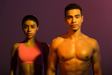 Fototapeta na wymiar attractive african american woman in pink sports bra and shirtless mixed race man looking at camera on purple background