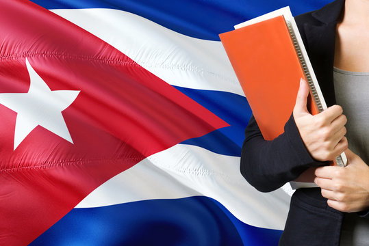 Learning Cuban language concept. Young woman standing with the Cuba flag in the background. Teacher holding books, orange blank book cover.