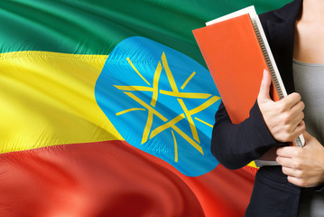 Learning Ethiopian language concept. Young woman standing with the Ethiopia flag in the background. Teacher holding books, orange blank book cover.