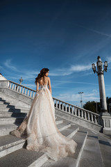 Obraz premium Beautiful model woman with perfect body in luxury wedding dress stands with back on the stairs and posing with blue sky at background