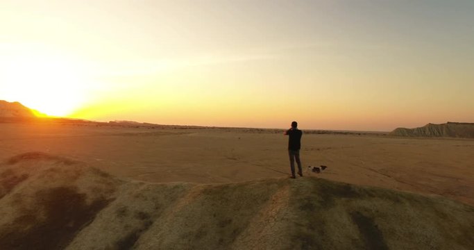 A rotating aerial shot of a man and his dog playing harmonica during sunset An aerial shot of a man and his puppy standing on top of a hill  and playing harmonica long shadows golden hour