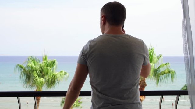 Young Man Opening Curtains And Going Out Onto Balcony To Enjoy View. Male On Vacation Looking At Mediterranean Sea From Terrace And Using Mobile Phone. Guy Talking To Friend Via Cellphone