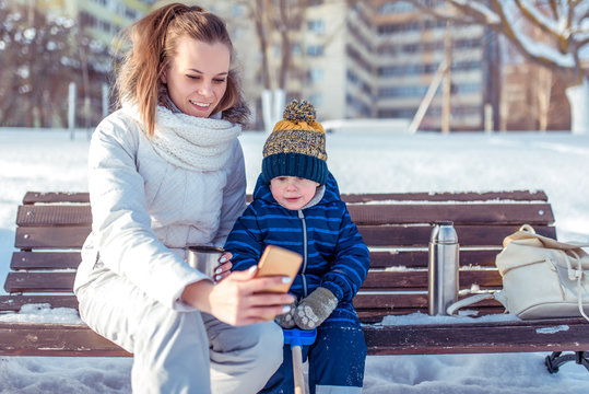 Young woman with a little son boy of 4 years. Talking on a video call. Sits in winter on a bench background of snow and drifts. Online communication on the Internet, take pictures of themselves.