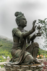 Hong Kong, China - March 7, 2019: Lantau Island. Side Closeup, One of the Six Devas offers lamp to Tian Tan Buddha. Bronze statue seen from front with green foliage and rainy sky in back.