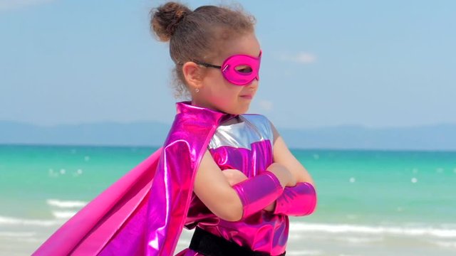 Beautiful Little Girl in the Superhero Costume, Dressed in a Red Cloak and the Mask of the Hero. Plays on the Background Sea and Blue Sky and Clouds, Sends a Fist Forward. Concept of a Happy Chilhood.