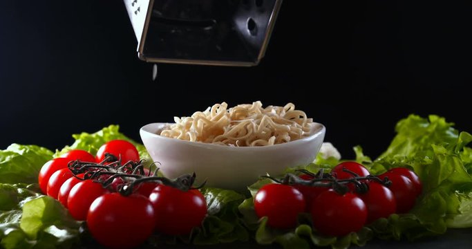 On a black background, vegetables and noodles on a wooden table, the whole dish is rubbed on top with parmesan cheese and mixed noodles. Concept of: Grater, Cheese, Noodles, Cherie Tomatoes, Salad.