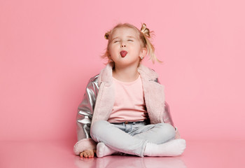 Portrait of a little girl in stylish clothing sitting on pink background and playing up