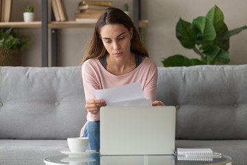 Serious woman sitting on couch reading letter feels frustrated
