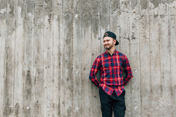 bearded man in a plaid shirt leaning against a gray wall standing in the open air