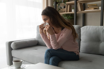 Unhealthy woman sitting on couch holding tissue wiping her nose