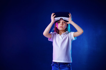 Girl 7 y.o. experiencing VR headset game on colorful background. Child using a gaming gadget for virtual reality. Picked it up and looking at camera questionally. Virtual technology