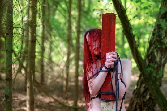 young sexy naked woman with red hair is tied to a post with a rope in the woods. 