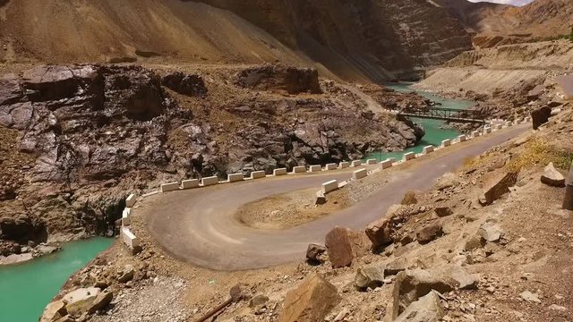 Wide shot of orange vans on a mountain road, switchbacking down a steep canyon slope, and eventually crossing the canyon's river at a bridge