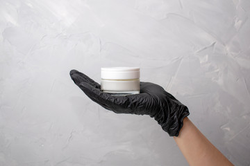 Female hands in black medical glove holds jar with cream. Women's hands with white cosmetic jar on the grey background.