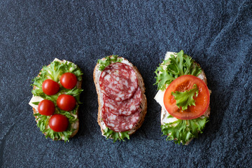 Fresh sandwiches with sausage, cheese, bacon, tomatoes, lettuce, cucumbers on a dark background. Horizontal orientation