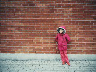 Portrait of smiling cute adorable little preschool girl in pink winter jacket standing leaning on red brick wall outside in street and looking away waiting for somebody
