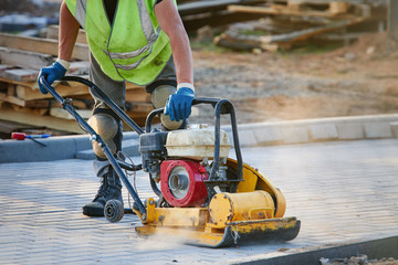 Worker in uniform and knee pads use vibratory plate compactor for path construction. Plate compactor for compaction soil or pavement or sidewalk. Indastrial equipment. Laying and tamping paving slabs