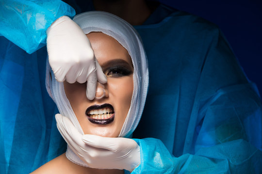 Doctor In Hygiene Suit Gloves Stand Back Of Patient, Twitching Nose Reshaping Treatment Or Rhinoplasty On Asian Woman With Bandage After Plastic Surgery Operating. Studio Lighting Blue Background