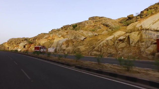 Handheld, tracking, exterior shot inside car driving alongside rocky hills and trucks.