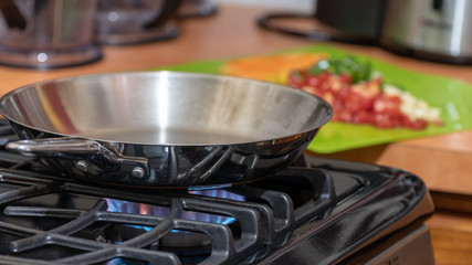 Stainless steel frying pan on lit modern gas stove in preparation for cooking food. Chopped ingredients for meal blurred in background on green chopping/cutting board on counter top. 