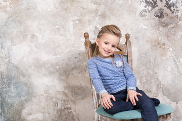 Portrait of a little attractive smiling girl in a blue sweater and pants with her hair folded in her hair sitting on a chair against a grunge wall background.