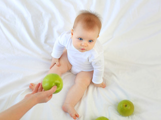 Fototapeta na wymiar Charming blue-eyed 9-month-old kid with funny hair sitting on the bed with green apples. White clothes, blond hair.
