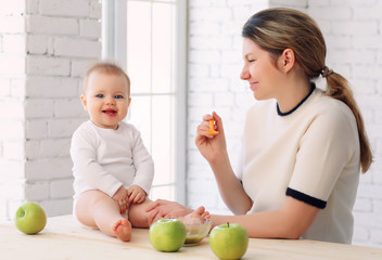 Fototapeta premium 9 month old charming baby sits on the table and mom feeds him with a spoon of applesauce. Blonde hair, blue eyes.