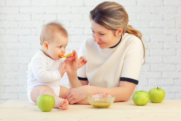 Fototapeta premium 9 month old charming baby sits on the table and mom feeds him with a spoon of applesauce. Blonde hair, blue eyes.